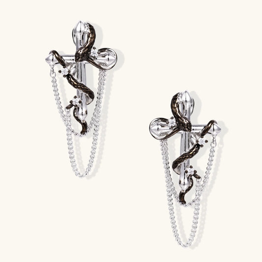 Silver dangle earrings with sculptural serpent coiled around cross shape and delicate chain accents on cream background