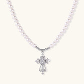 White pearl necklace with ornate silver cross pendant featuring zirconia crystals on cream background.