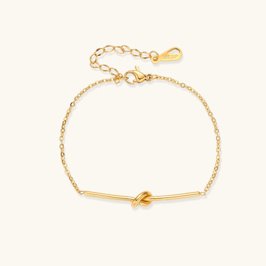 Delicate gold chain bracelet with minimalist bar and centered knot detail on cream background