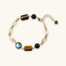 Gold chain bracelet with tiger's eye, obsidian, pearl, and iridescent abalone stones on cream background.