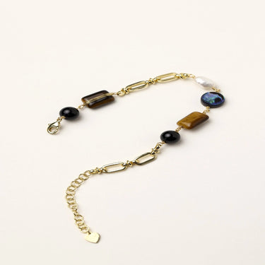 Gold chain bracelet with tiger's eye, obsidian, pearl, and iridescent abalone stones on cream background.